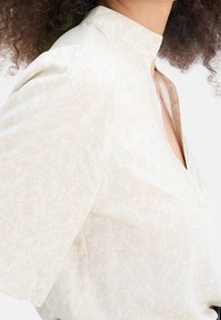 Krémová blůza s jemným vzorem, stojáčkovým límečkem a volnými, krátkými rukávy. Lehká tkanina s hladkou strukturou.