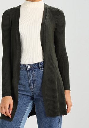 Kobieta w długim, ciemnozielonym, prążkowanym kardiganie na białym prążkowanym golfie oraz niebieskich spodniach z wysokim stanem na jednolitym tle.