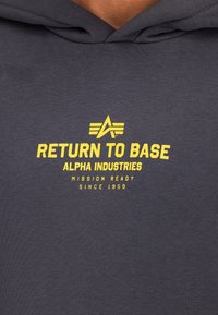 Harmaa huppari, jossa keltaiset tekstit: "RETURN TO BASE", "ALPHA INDUSTRIES", "MISSION READY" ja "SINCE 1959." Pehmeää puuvillakangasta.