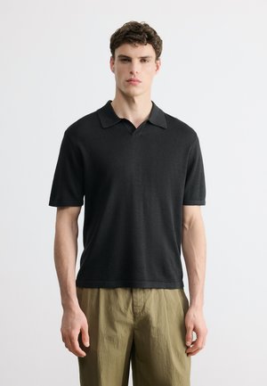 Jeune homme aux cheveux courts et bouclés, portant une chemise noire à manches courtes avec col et un pantalon vert olive, debout devant un fond blanc uni.