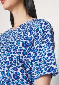 Femme aux cheveux lisses et foncés portant un haut à manches courtes imprimé léopard bleu et blanc sur un fond clair uni.