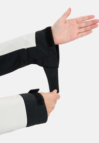 Czarna i biała rękaw kurtki z zapięciem na rzep; gładka tekstura, sportowy design; ręka pokazująca mechanizm mocowania dla regulowanego dopasowania.