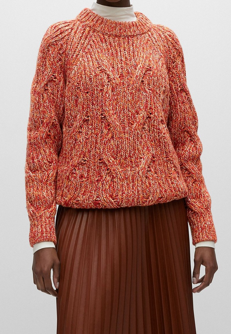 Gestrickter Pullover in Rot- und Orangetönen mit grober Textur, weitem Rundhalsausschnitt und gerippten Bündchen. Kombiniert mit einem braunen Faltenrock.