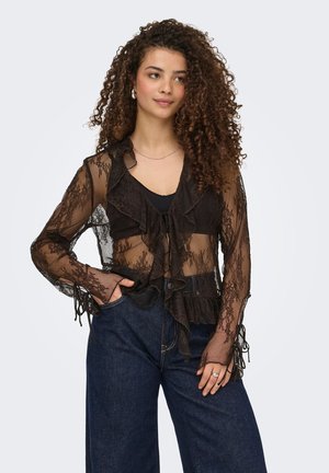 Femme aux cheveux bouclés portant un chemisier en dentelle noire transparente sur un haut noir et un jean bleu foncé taille haute, debout avec une main dans la poche.