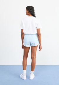 T-shirt blanc court associé à un short élastique bleu clair. Le modèle porte des baskets et des chaussettes blanches, avec des cheveux tressés. Fond uni.