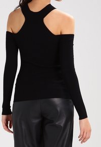 Frau trägt ein schwarzes, kalt-schulteriges, geripptes Stricktop und schwarze Lederhosen, von hinten vor plain Hintergrund gezeigt.