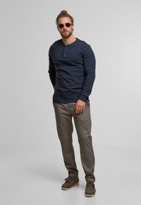 Camicia henley a maniche lunghe blu navy con tre bottoni, pantaloni a scacchi grigi e scarpe mocassino marroni, indossati con occhiali da sole rotondi.