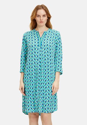Türkises Tunika-Kleid mit geometrischem Muster in Grün und Blau. Lange Ärmel und ein V-Ausschnitt mit kontrastierenden Besatzdetails. Aus weichem Stoff.