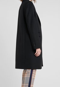 Person som bär en svart knälång kappa över rutiga byxor med marinblå och orangea sidostråk, stående mot en enkel ljus bakgrund.