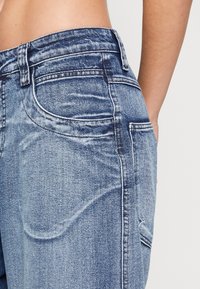 Blå denimjeans med hög midja, tydlig sömnad, rundade fickor och en urtvättad finish, som visar upp ett texturerat, lätt material.