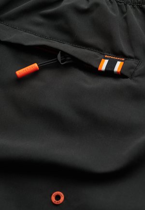 Black fabric with a smooth texture, featuring an orange drawstring, zip pocket, and a small Superdry tag in black, orange, and white stripes.