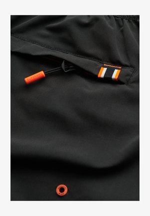 Black fabric with a smooth texture, featuring an orange drawstring, zip pocket, and a small Superdry tag in black, orange, and white stripes.