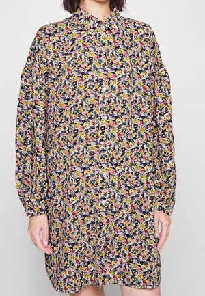 Femme portant une robe fleurie ample à manches longues avec des fleurs roses, jaunes, blanches et bleu foncé, et une patte de boutonnage à l'avant.