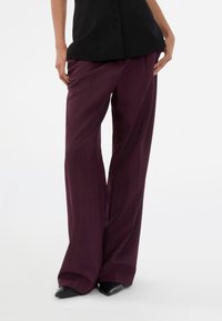 Wide-leg trousers in deep burgundy fabric, featuring a high waist and smooth texture. Paired with a black top, held with one hand.