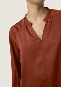 Blusa color ruggine in tessuto liscio, con scollatura a V, maniche lunghe e una sottile texture con dettagli arricciati sulle spalle.