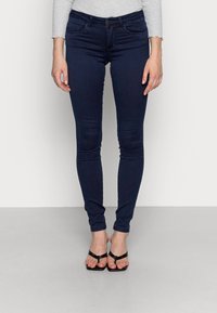 Skinny dark blue jeans made of denim, featuring a mid-rise waist, five pockets, and a button closure. Worn with black flip-flops.
