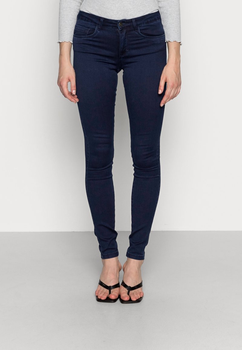 Skinny dark blue jeans made of denim, featuring a mid-rise waist, five pockets, and a button closure. Worn with black flip-flops.
