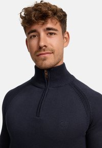 Granatowy sweter zapinany na zamek z dzianiny, z prążkowanym kołnierzem, teksturowymi detalami na ramionach i małym logo na klatce piersiowej. Miły w dotyku, dopasowany krój.