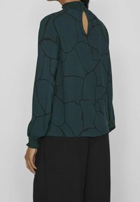 Blusa verde a fantasia con maniche lunghe, colletto arricciato e dettaglio a occhiello sul retro. Presenta linee astratte nere su un tessuto morbido.