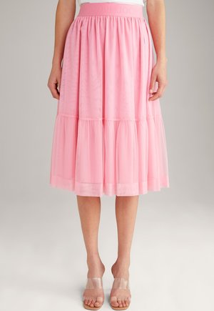 Femme portant une jupe midi rose à volants avec une large ceinture et une superposition transparente, associée à des talons ouverts à brides transparentes.
