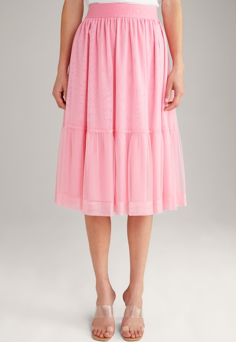 Femme portant une jupe midi rose à volants avec une large ceinture et une superposition transparente, associée à des talons ouverts à brides transparentes.