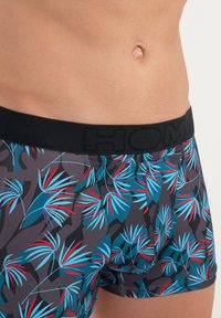 Herren-Boxershorts mit einem tropischen Blättermuster. Sie verfügen über einen schwarzen elastischen Bund mit "HOM"-Branding und lebhaften blauen und roten Blättern auf einem dunklen Hintergrund.