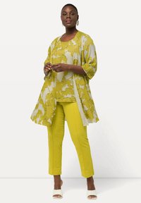 Kimono fluido e trasparente giallo con motivo floreale su una maglia gialla solidale e pantaloni aderenti. Tacchi aperti bianchi completano il look.