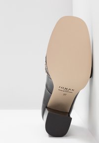 Chaussure en cuir noire à talon, posée sur son talon avec la semelle tournée vers l'avant montrant la marque Jonak et la taille 37.