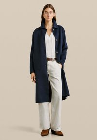 Marineblauwe wollen jas met een kraag en knoopsluiting, gecombineerd met een witte top en wijde witte broek. Model staat tegen een neutrale achtergrond.