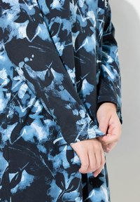 Indumento a manica lunga di un blu scuro con motivo floreale blu; tessuto morbido con una texture liscia; mano che regola il polsino della manica.