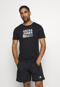 Black cotton T-shirt with "JACK & JONES PERFORMANCE" graphic in white. Paired with black shorts featuring an Adidas logo on the hem.