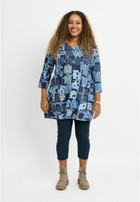 Marineblaues Tunika mit einem Patchwork-Muster in Blau- und Grüntönen, kombiniert mit dunklen Leggings und braunen Knöchelstiefeln. Freizeitliche 3/4-Ärmel.