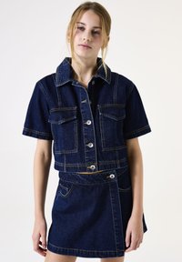 Donker denim korte mouwen jasje met twee borstzakken, zilveren knopen en gele stiksels. Aangesloten stijl met een bijpassende korte rok.