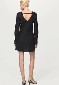 Mujer de espaldas llevando un mini vestido negro de manga larga con un escote en V profundo y tirantes finos, combinado con sandalias negras de tacón.