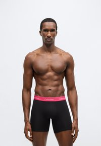 Chiloți boxer negri cu talie roz Calvin Klein, având o croială strâmtă și o textură moale. Modelul pozează frontal, evidențiind articolul vestimentar.