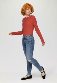 Haut à manches longues en ribbed rouille, jeans slim bleu avec déchirures au genou, et ballerines noires avec une sangle. Le mannequin est dans une pose dynamique.