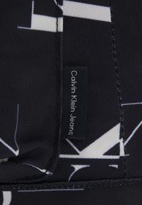 Etichetta Calvin Klein Jeans su un materiale nero testurizzato con un motivo geometrico bianco e dettagli di cucitura a contrasto.