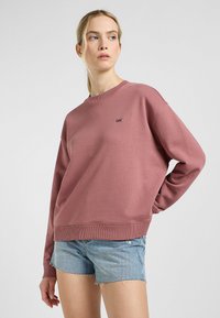 Sudadera rosa con puños y dobladillo acanalados, que presenta un pequeño logo negro en el pecho. Combinada con pantalones cortos de mezclilla azul claro.