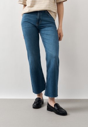 Persoon met blauwe cropped flare jeans met rafelige zomen en zwarte leren loafers, staand tegen een effen lichte achtergrond.