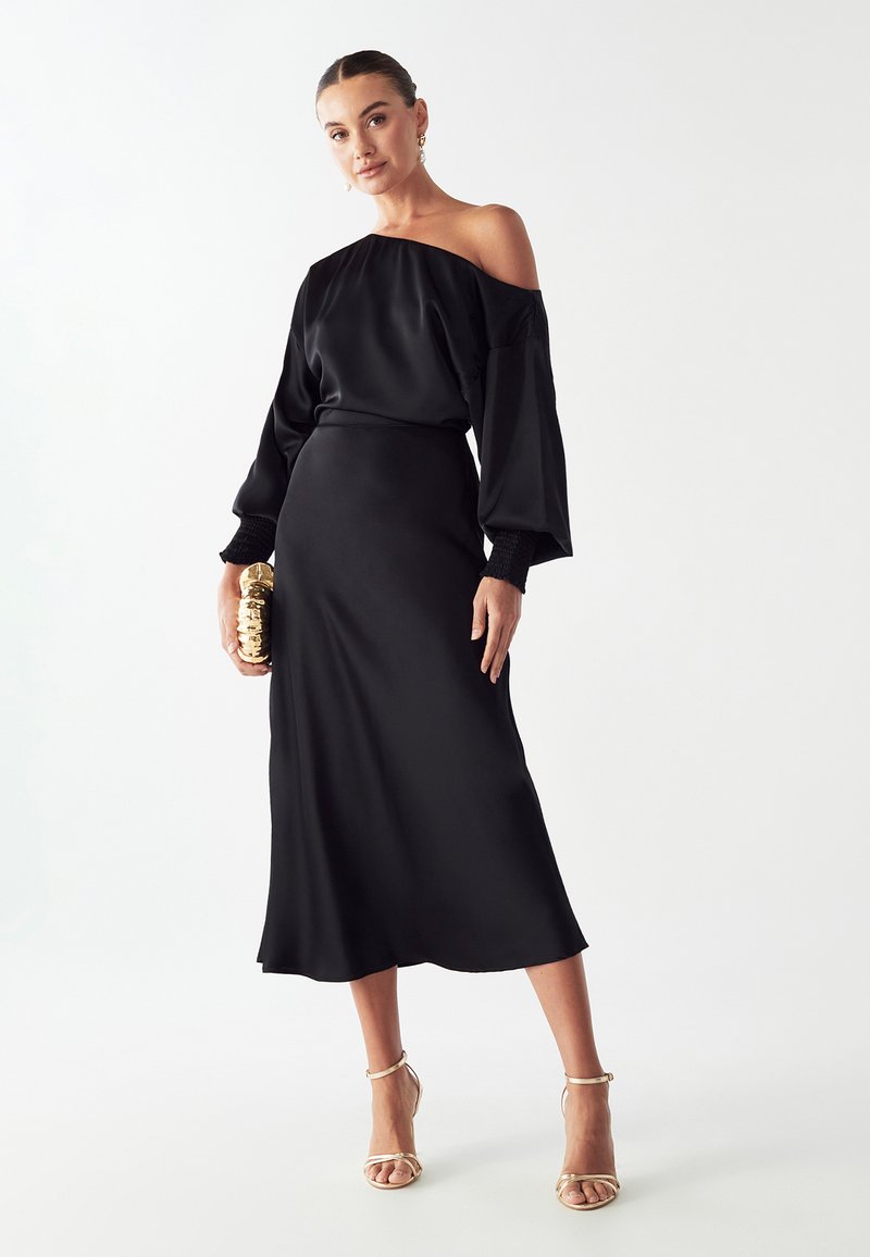Čierne saténové šaty bez ramienok s dlhými balónovými rukávmi, elastickými detailmi na manžetách a splývavou sukňou do polovice lýtok. Zlaté púzdro ako doplnok.