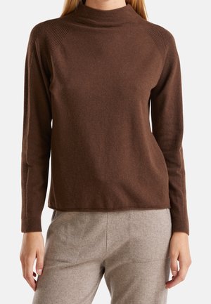 Femme portant un pull à col roulé marron à manches longues et un pantalon gris clair, debout avec les mains détendues le long du corps.