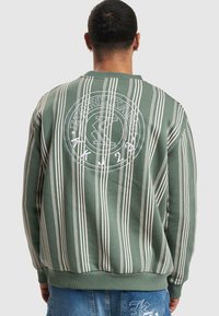Maglione verde a righe con linee verticali bianche e un logo circolare sul retro, dotato di polsini e orlo a coste. Tessuto casual e texturizzato.