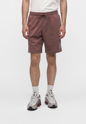 Homme portant un short marron à cordon, des chaussettes blanches et des chaussures de sport blanc-gris, debout devant un fond uni.