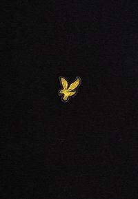 Tissu noir avec une texture lisse présentant un petit emblème d'aigle jaune brodé sur le côté supérieur gauche.