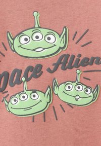 Grafika prikazuje tri zelena tuja glava s tremi očmi in šiljastimi ušesi na rožnati podlagi, ob njej je besedilo "Vesoljski tujci" v črni barvi.