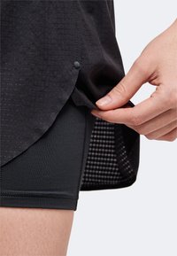 Main remontant l'ourlet d'un t-shirt de sport noir texturé, montrant un short noir ajusté en dessous sur un fond clair.