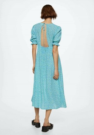 Femme portant une robe midi bleue à fleurs avec des manches bouffantes et un dos ouvert noué au cou, debout devant un fond uni.
