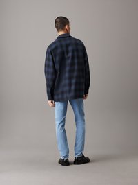 Camisa de cuadros azul marino con un corte relajado, mangas largas y cuello. Combinada con jeans azul claro y zapatos negros, vista desde la espalda.