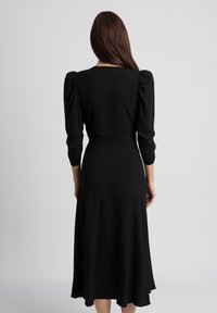 Kobieta z długimi brązowymi włosami, ubrana w czarną midi sukienkę z bufiastymi długimi rękawami, widziana z tyłu na jednolitym tle.