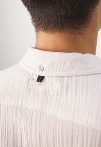Arrière du cou d’une personne aux cheveux courts et foncés portant une chemise blanche texturée boutonnée au col avec une boucle en tissu noire en dessous.